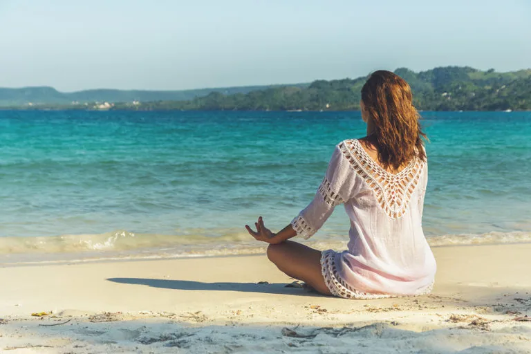Woman in white crocheted cover-up sitting in yoga pose on sandy beach overlooking turquoise ocean and forested coastline