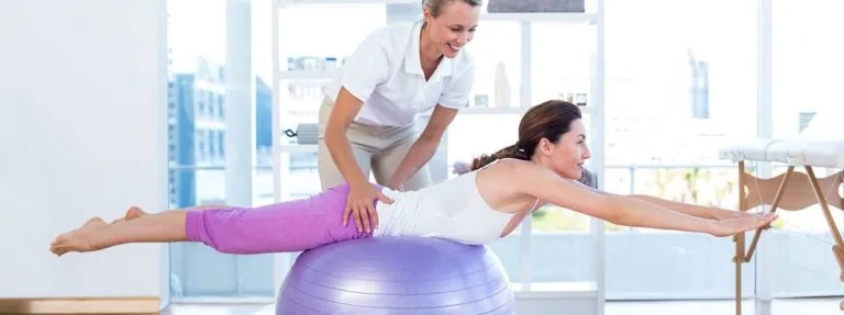Woman balancing on purple exercise ball with trainer providing instruction in bright fitness studio with city skyline view
