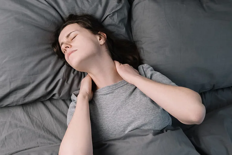 Woman lying on gray bedding with hand on neck, eyes closed in discomfort