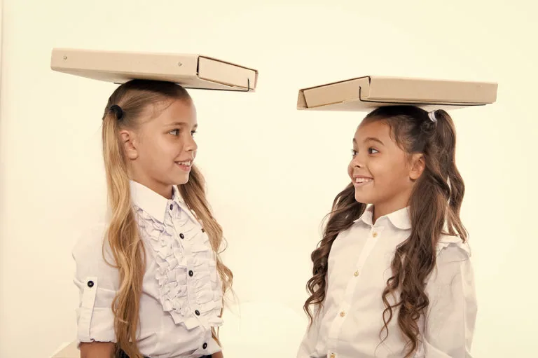 Two schoolgirls balancing cardboard boxes on their heads while looking at each other smiling
