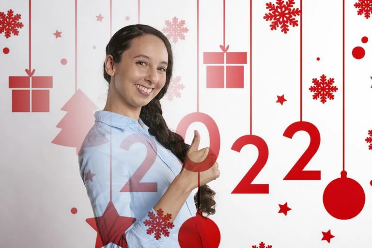 Girl smiling at camera with long dark hair wearing light blue shirt, standing in front of festive Christmas backdrop with hanging ornaments, snowflakes, and gift decorations