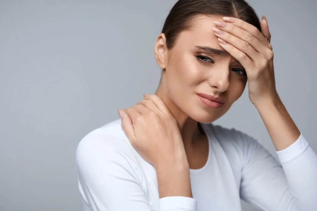 Woman in white shirt holding forehead and neck, expressing discomfort or pain