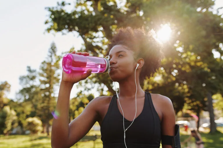 Woman drinking from pink water bottle outdoors during exercise with earbuds in