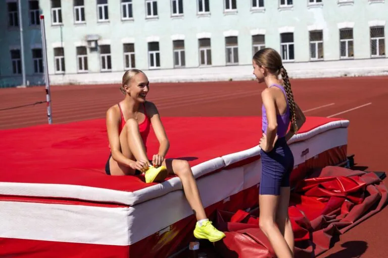 Two teenage girls in athletic wear sitting on a red gymnastics mat at a track facility, having a conversation