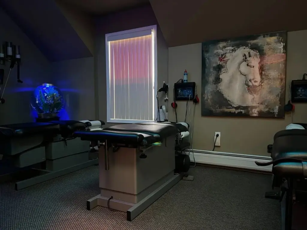 An adjusting room lit in soft blue: two padded chiropractic tables, a glowing orb lamp in the corner and a large horse painting on the wall