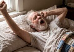 Older man stretching in bed with one arm raised above his head while yawning
